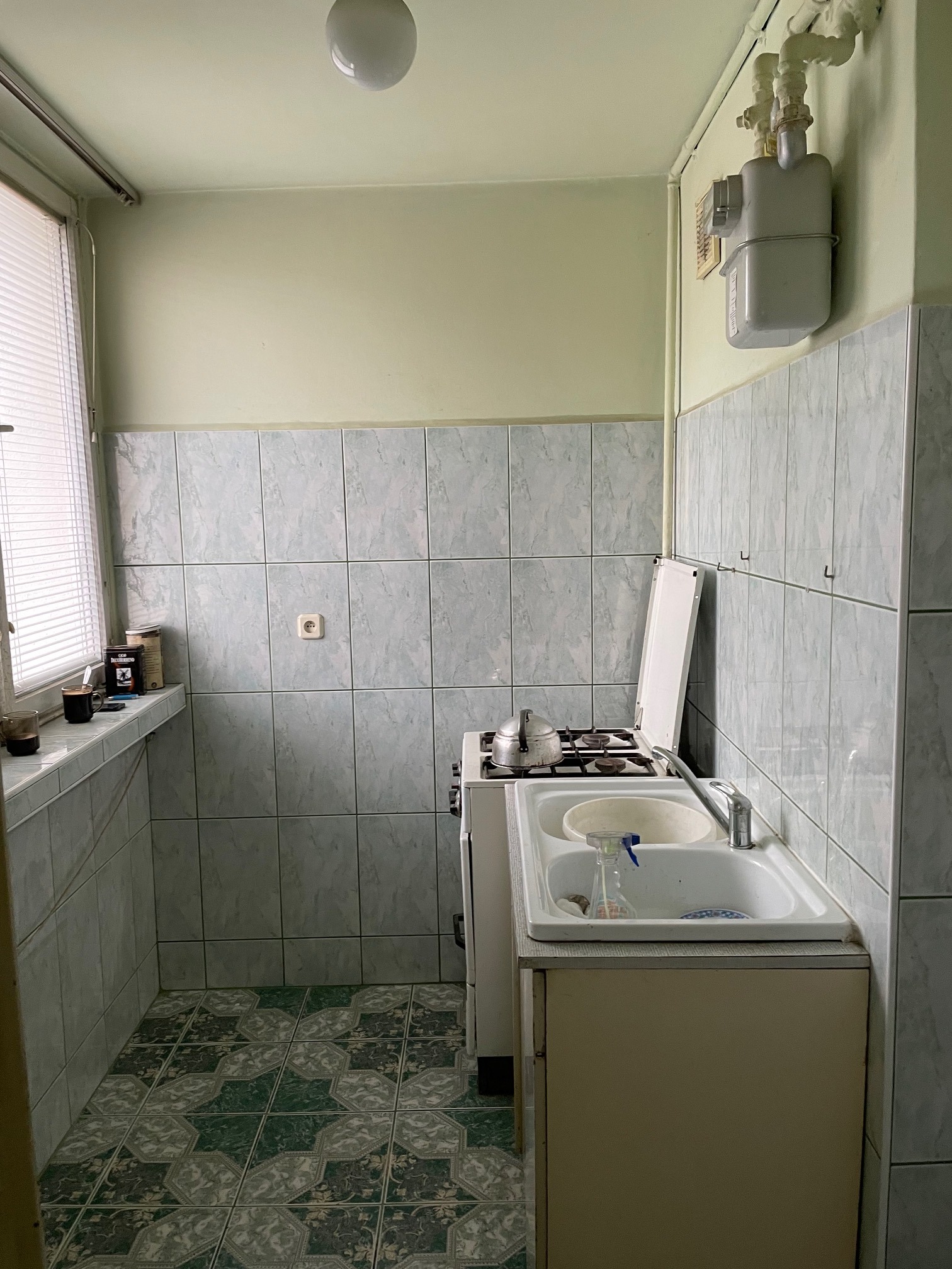 Mała, zaniedbana kuchnia z kuchenką gazową, zlewem, płytkami w kolorze seledynowym i zieloną podłogą z wzorem. Na blacie przy oknie stoją puszki i kubki.