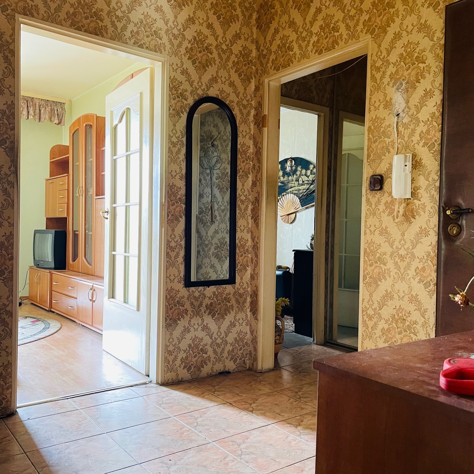 Wnętrze mieszkania z widokiem na kilka pomieszczeń przez otwarte drzwi, z meblami w stylu retro, wzorzystą tapetą i lustrem na ścianie.