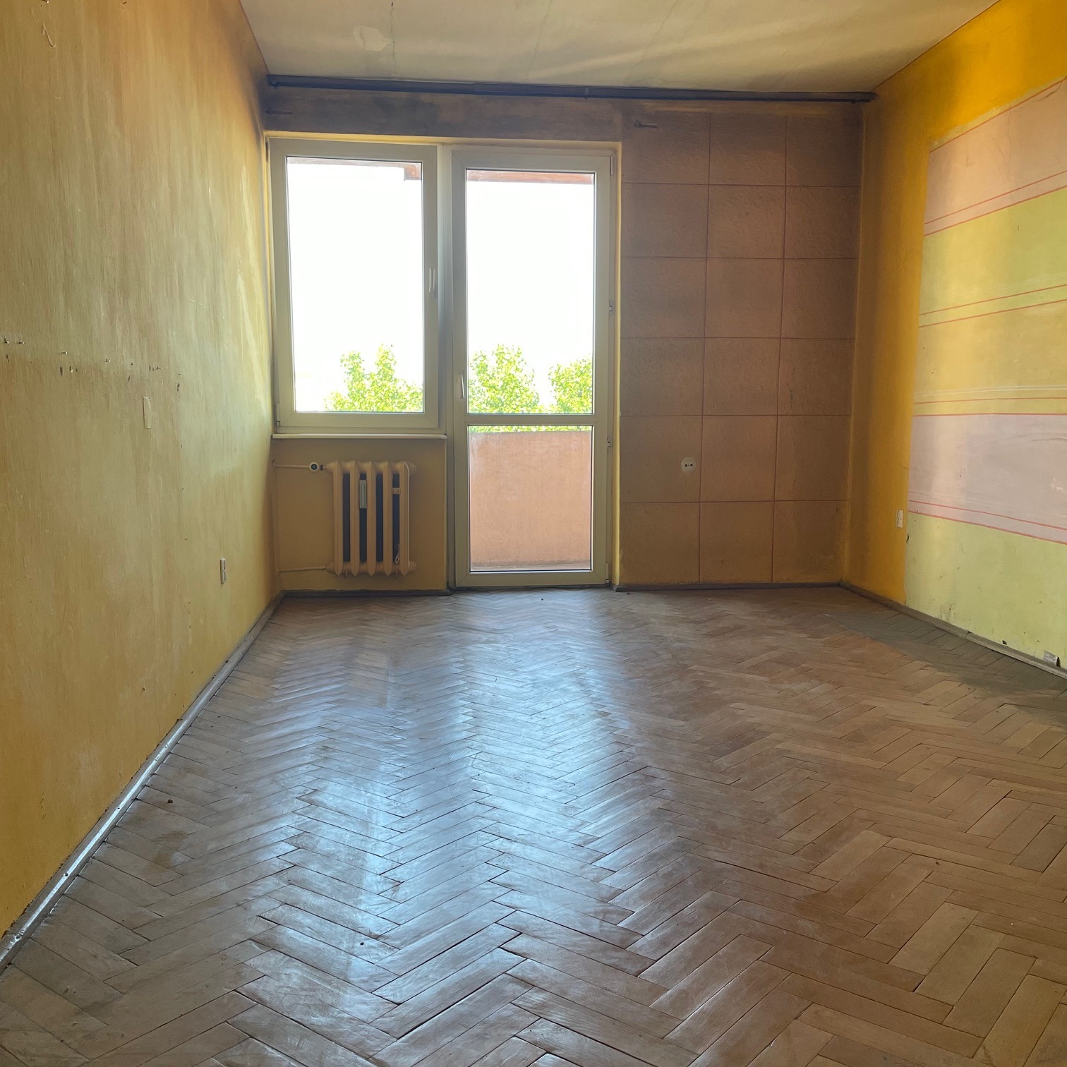 Pusty pokój przed remontem z parkietem w jodełkę, żółtymi ścianami, oknem balkonowym i grzejnikiem.