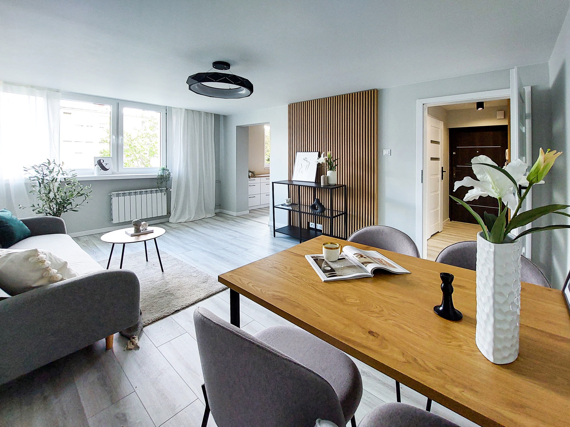 Jasne wnętrze mieszkania z drewnianym stołem, szarą sofą i dekoracyjną ścianką z lameli; nowoczesny design z akcentami roślinnymi i minimalistycznymi dodatkami.