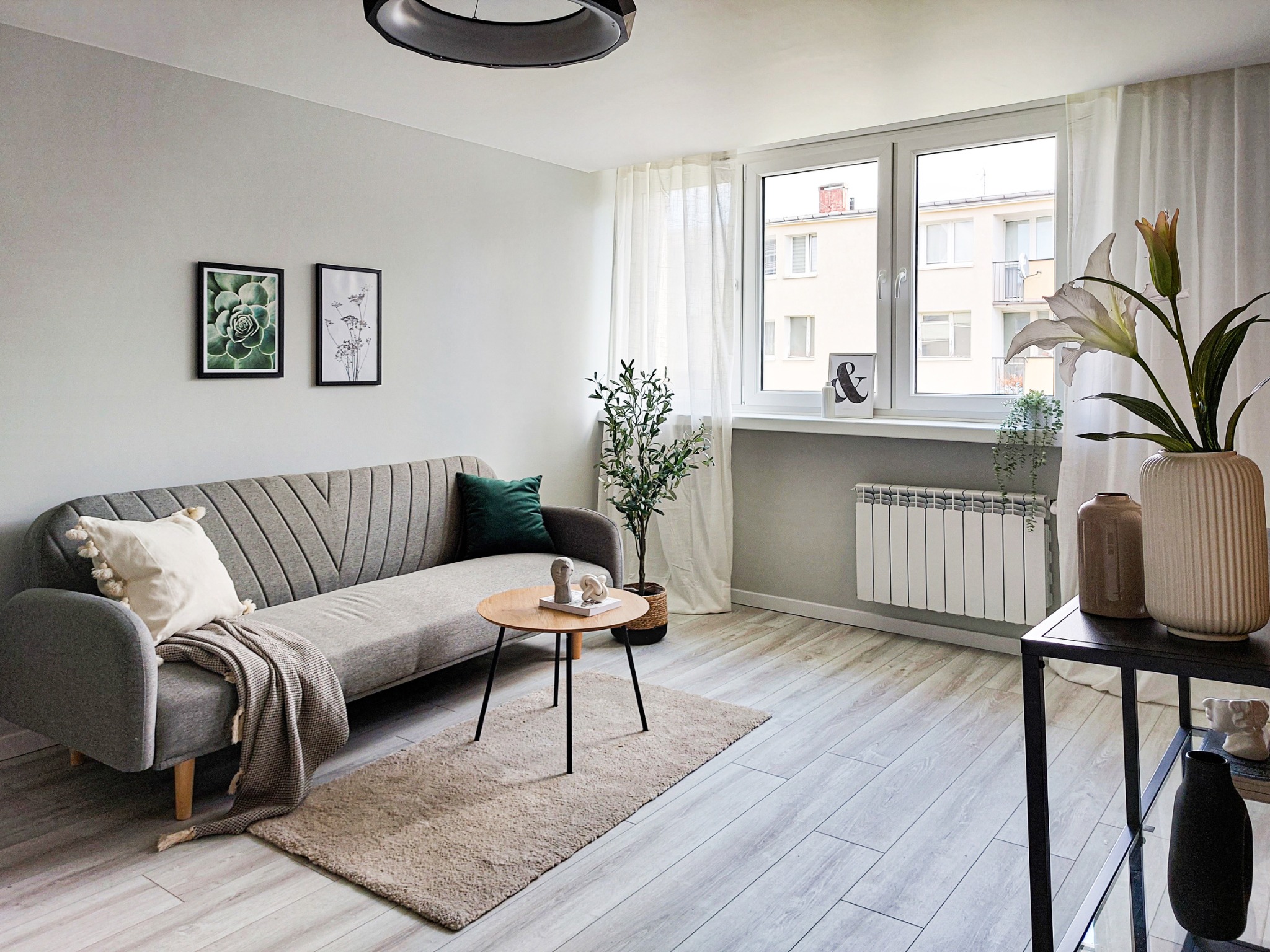 Jasne, minimalistyczne wnętrze salonu z szarą sofą, drewnianym stolikiem kawowym, dywanem, roślinami w doniczkach, obrazami na ścianie i dużym oknem z białymi zasłonami.