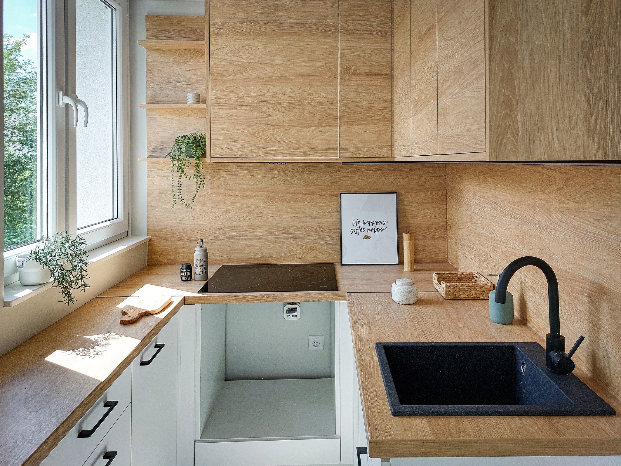 Nowoczesna kuchnia z drewnianymi frontami szafek, czarnym zlewem granitowym i indukcyjną płytą grzewczą, oświetlona naturalnym światłem wpadającym przez okno z zielonymi roślinami.