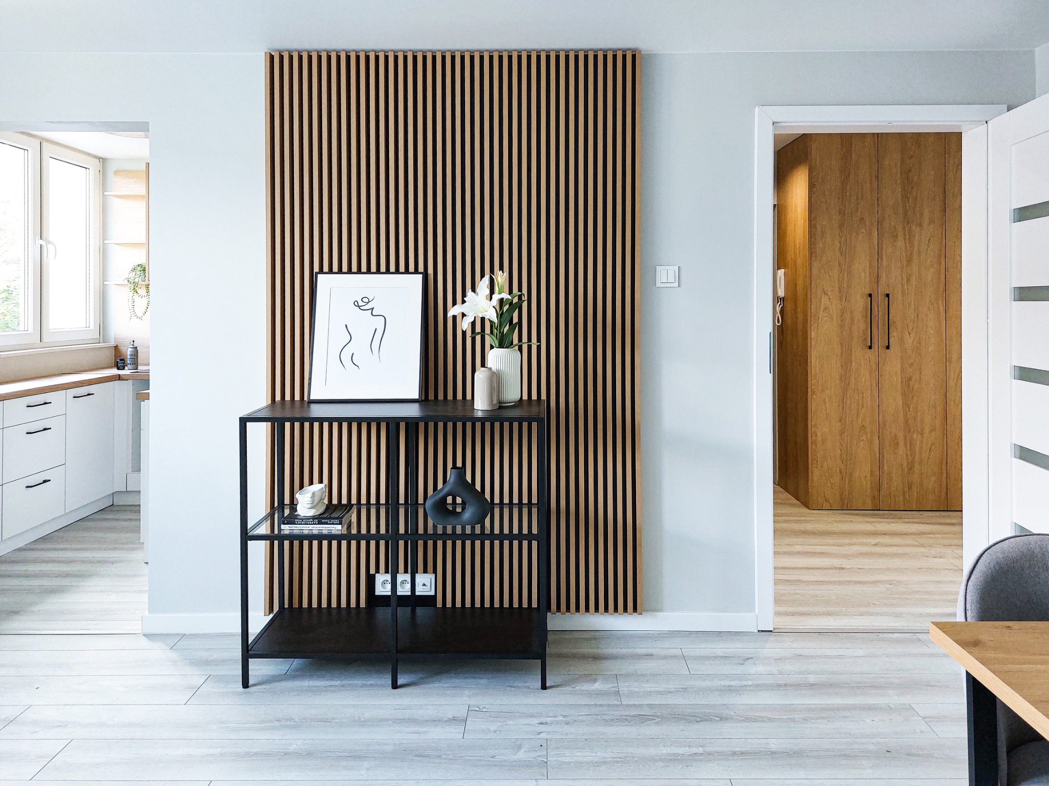 Nowoczesne wnętrze mieszkania z drewnianą ścianką dekoracyjną z pionowych listew, czarnym metalowym regałem z dekoracjami, minimalistycznym obrazem i białymi liliami w wazonie, widok na kuchnię...