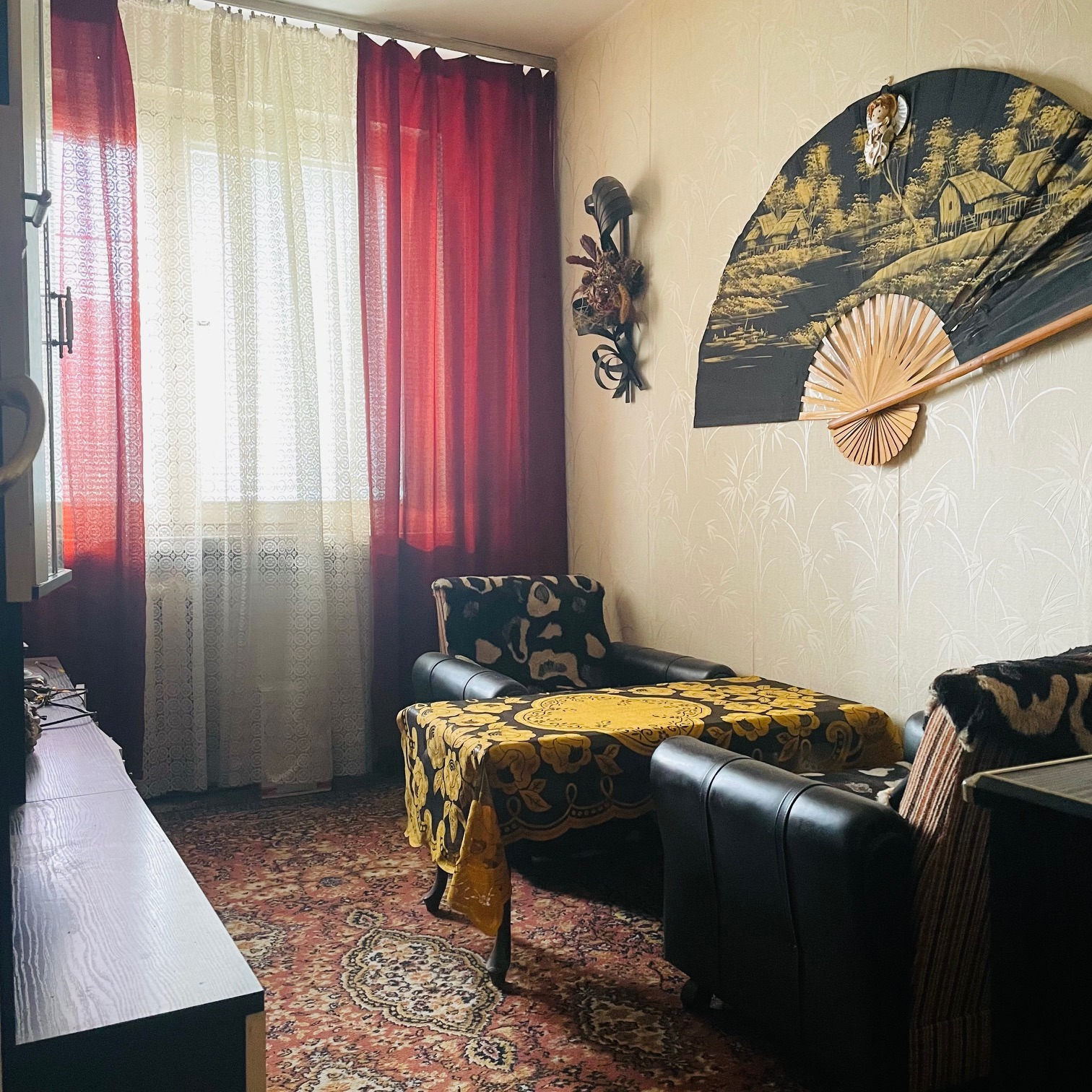 Aranżacja pokoju z ciemnymi meblami, orientalnym wachlarzem na ścianie i wzorzystym dywanem, oświetlona światłem dziennym wpadającym przez okno z czerwono-białymi zasłonami.