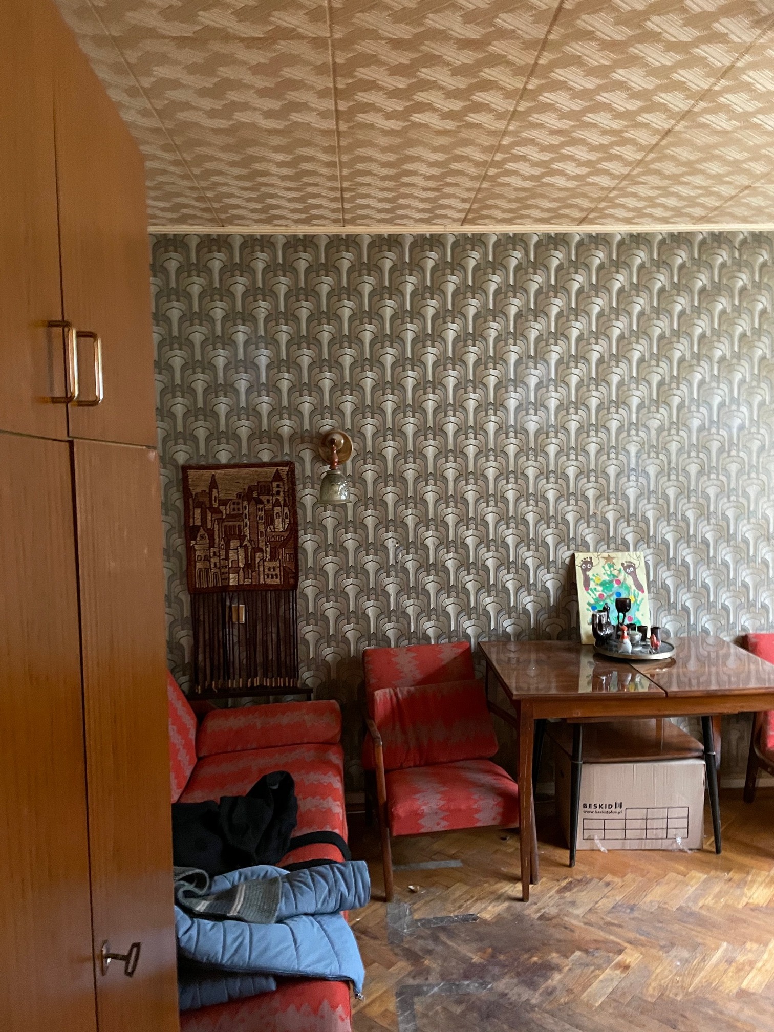 Pokój z meblami w stylu retro: wzorzysta tapeta, drewniany stół z dekoracją, czerwona sofa z ubraniami, szafa z metalowymi uchwytami i parkiet na podłodze.