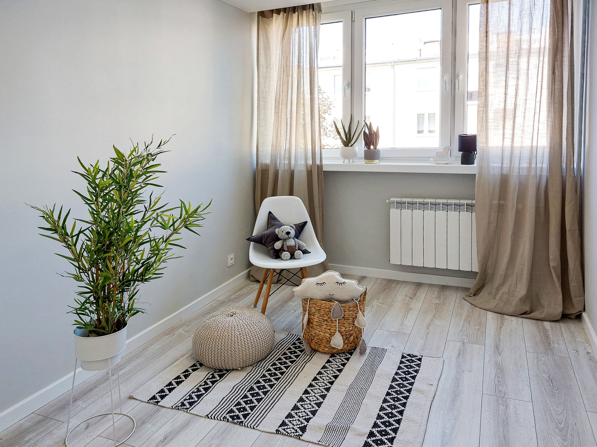 Jasne wnętrze pokoju z bambusową rośliną w białej donicy, dywanem w geometryczne wzory, pufem, krzesłem z pluszowym misiem i koszem z zabawką w kształcie chmury, oświetlone naturalnym światłem...