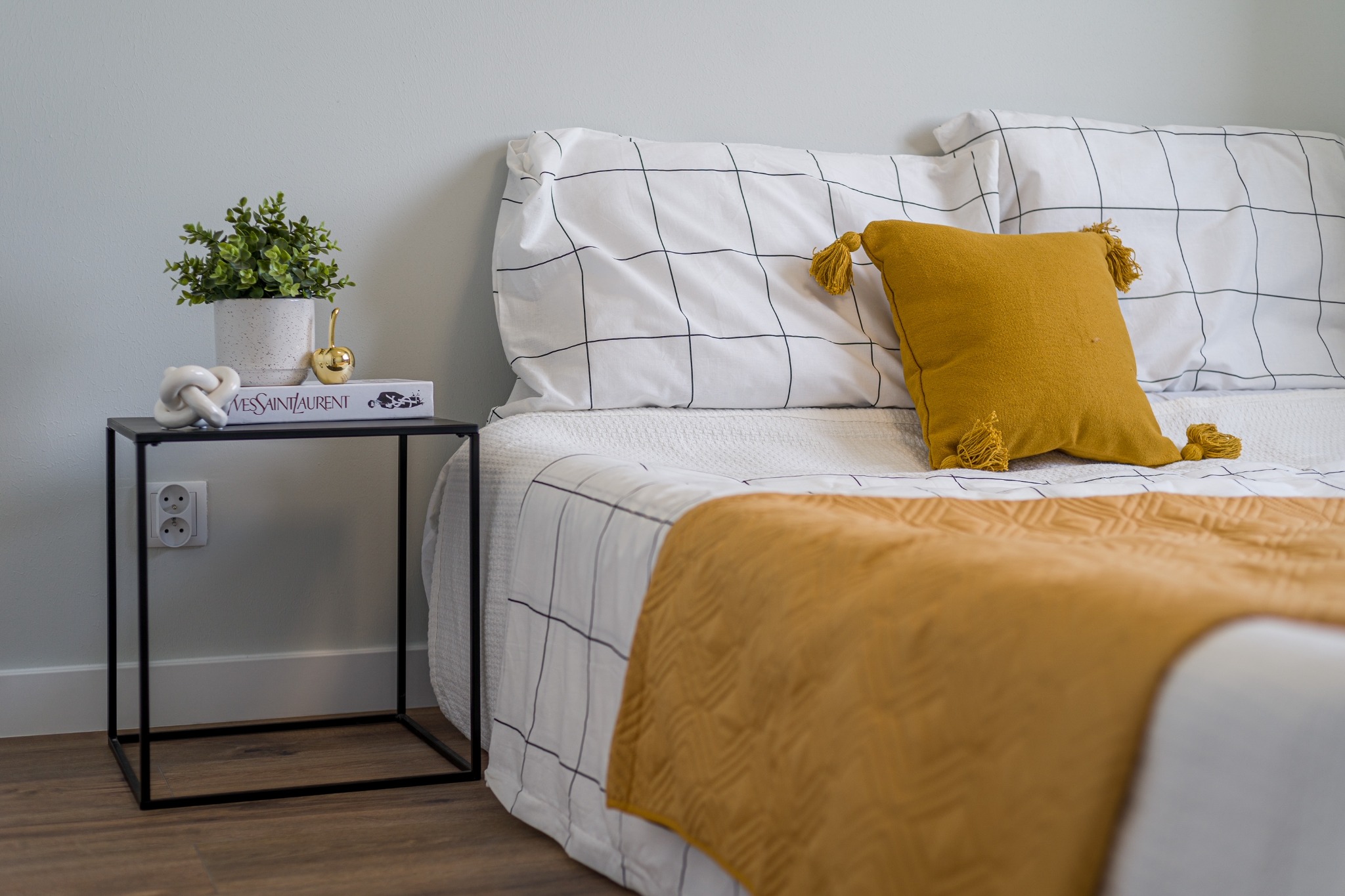 Sypialnia z łóżkiem w kratę, żółtą narzutą i poduszką z frędzlami, stolik nocny z książką YSL, rośliną i ozdobną kulką.