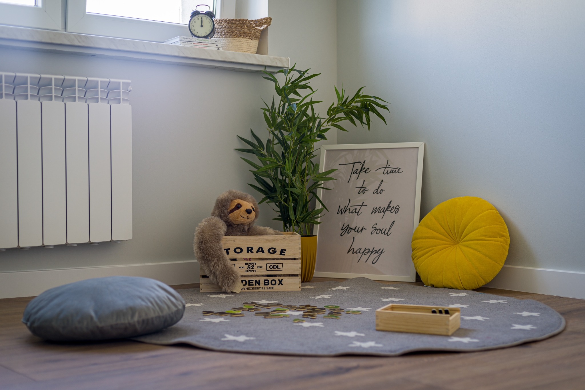 Pokój dziecięcy z okrągłym dywanem w gwiazdki, drewnianą skrzynką z pluszowym leniwcem, zieloną rośliną doniczkową, żółtą poduszką i plakatem z napisem 'Take time to do what makes your soul happy'.