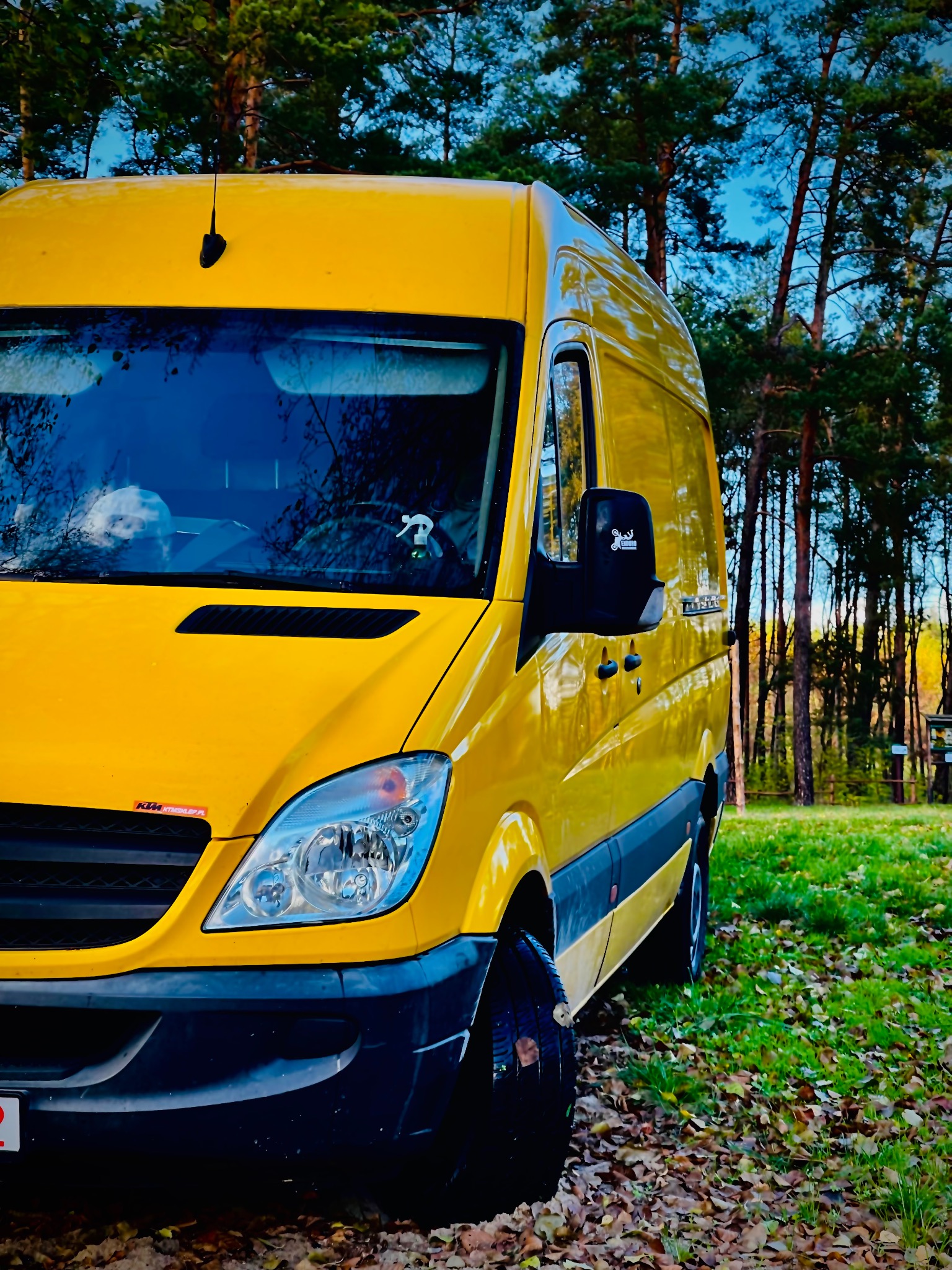 Żółty bus transportowy Mercedes-Benz Sprinter zaparkowany na trawie pokrytej liśćmi, widoczne logo firmy na lusterku i naklejka na masce, las w tle.