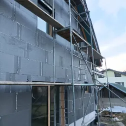 Dom w trakcie ocieplania styropianem grafitowym, widoczne rusztowanie, okna z drewnianą ramą i pianka uszczelniająca między płytami izolacyjnymi.