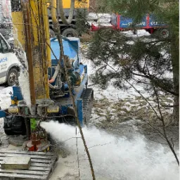 Wiercenie otworu w ziemi za pomocą specjalistycznego sprzętu, woda tryska z otworu pod ciśnieniem, widoczny pojazd ciężarowy i bus firmy w tle.