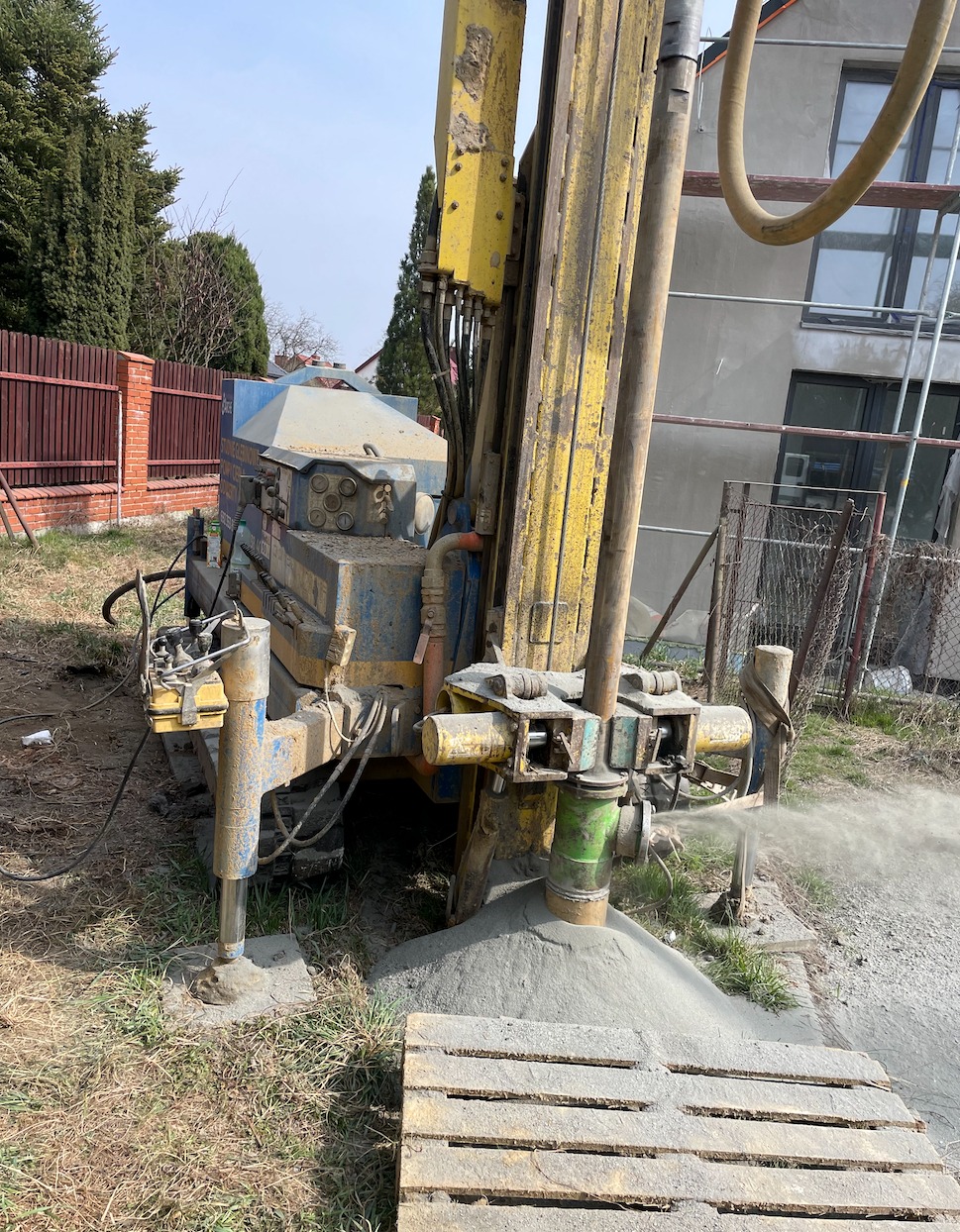 Wiertnica do studni w trakcie pracy, z widocznym wypływem wody i urobku, na tle domu w budowie i ogrodzenia.