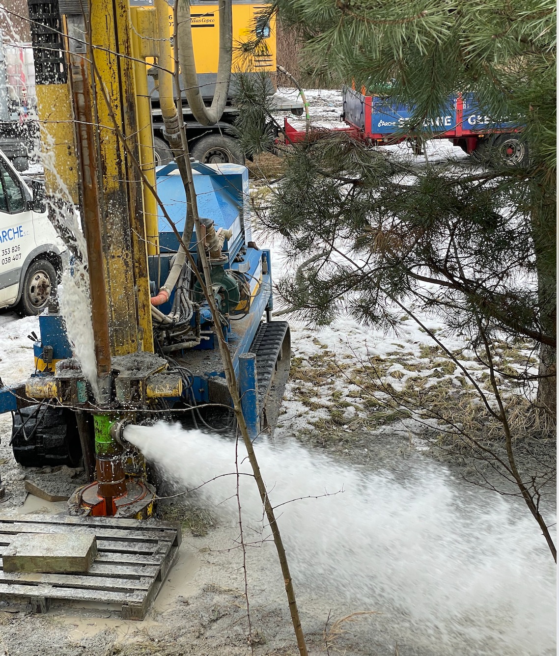 Wiercenie otworu w ziemi za pomocą specjalistycznego sprzętu, woda tryska z otworu pod ciśnieniem, widoczny pojazd ciężarowy i bus firmy w tle.