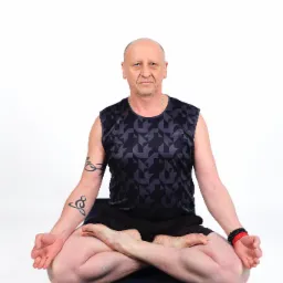 Mężczyzna w czarnym sportowym stroju siedzi w pozycji lotosu na ciemnej macie do ćwiczeń, z tatuażem na lewym ramieniu i bransoletką na prawym nadgarstku, na białym tle.