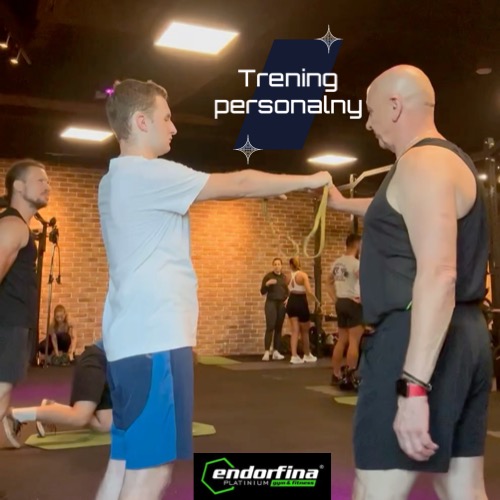 Mężczyzna w białej koszulce ćwiczy z gumą oporową pod okiem instruktora w siłowni Endorfina Platinium Gym & Fitness, na tle ceglanej ściany i innych ćwiczących.