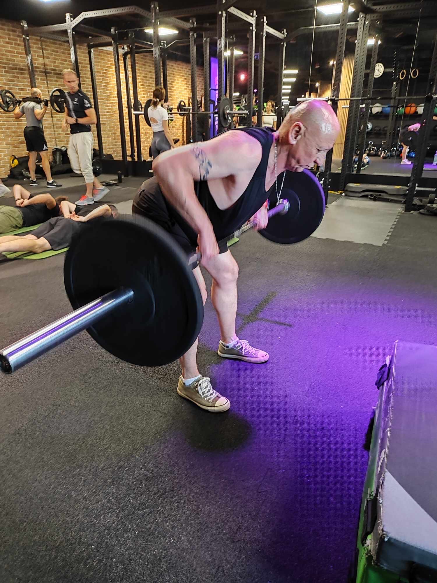 Mężczyzna w czarnym tank topie i krótkich spodenkach wykonuje wiosłowanie sztangą w opadzie tułowia w siłowni, na tle ceglana ściana i inni ćwiczący.