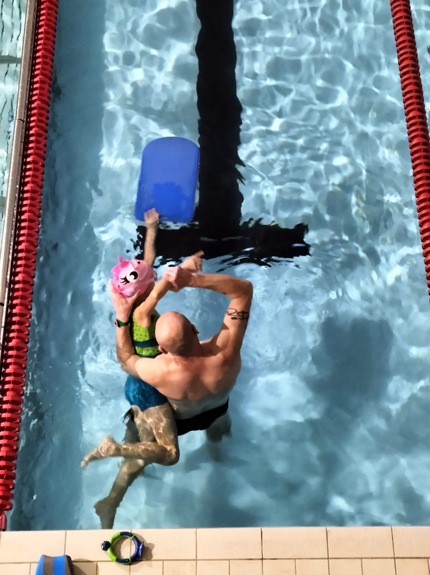 Instruktor z tatuażem na ramieniu uczy dziewczynkę w różowym czepku pływać w basenie, trzymając ją i niebieską deskę do pływania.