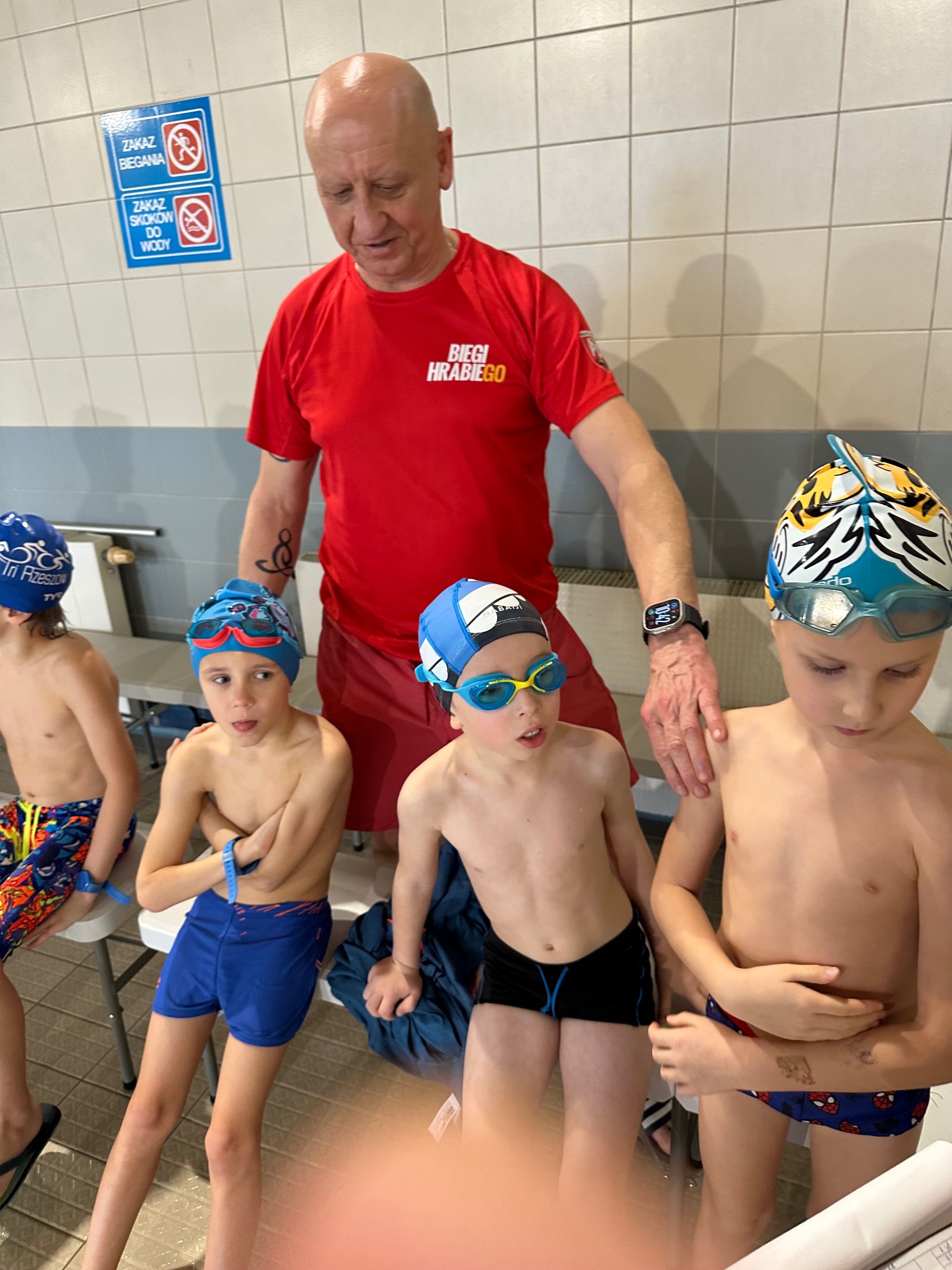 Instruktor w czerwonej koszulce z napisem 'Biegi Hrabiego' obok grupy chłopców w czepkach i okularach pływackich siedzących na ławeczce przy basenie, na ścianie tabliczka z zakazem biegania...