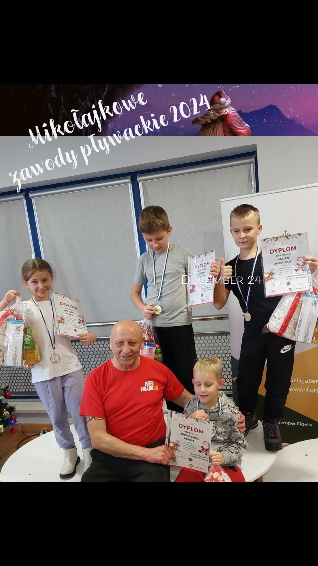 Dzieci z medalami i dyplomami na podium po Mikołajkowych zawodach pływackich 2024, z nagrodami w torbach, obok trener w czerwonej koszulce, na tle dekoracji świątecznych.