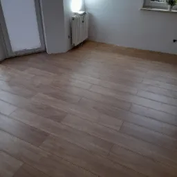 Ułożone panele podłogowe o wzorze drewna, widoczne przy ścianie z grzejnikiem i oknem z rośliną doniczkową.