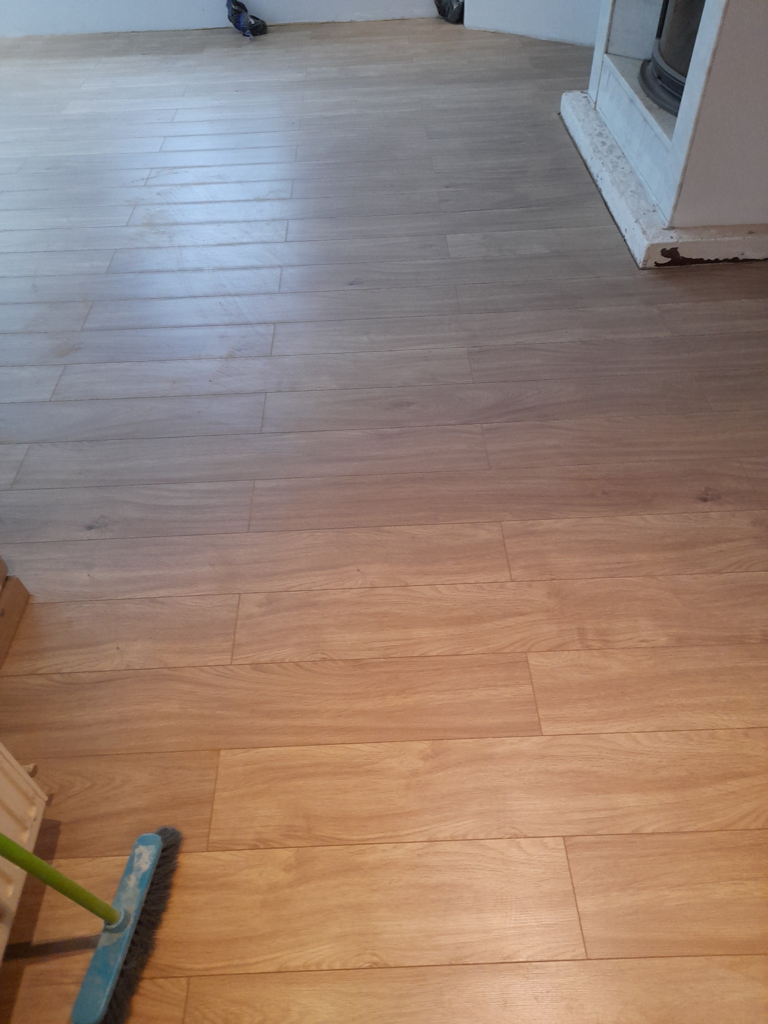 Ułożone panele podłogowe imitujące drewno, z widoczną szczotką do zamiatania w lewym dolnym rogu i fragmentem kominka w prawym górnym rogu, w tle widoczne ślady po remoncie.