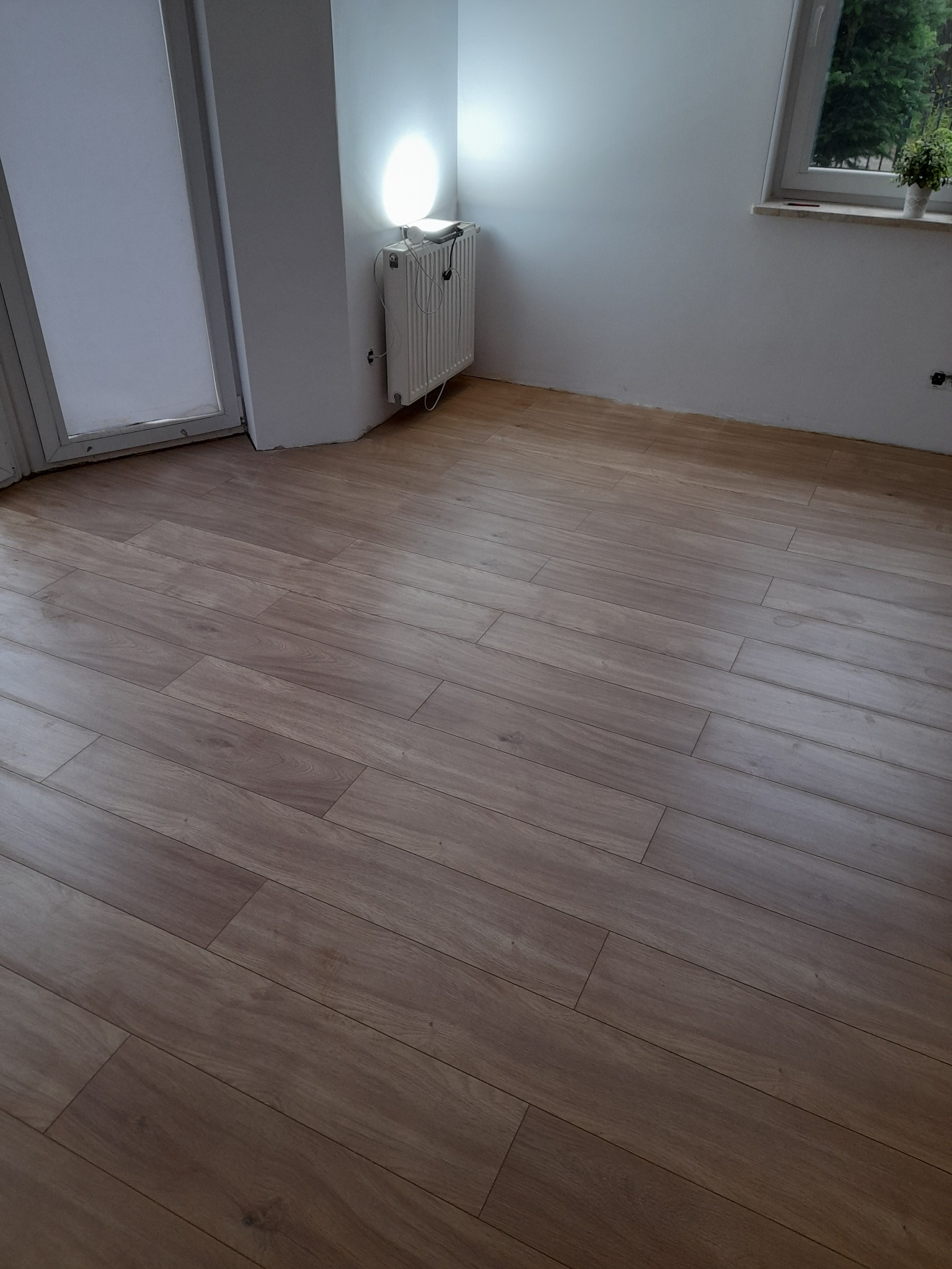 Ułożone panele podłogowe o wzorze drewna, widoczne przy ścianie z grzejnikiem i oknem z rośliną doniczkową.