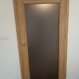 Zamknięte drzwi wewnętrzne z drewnianą ramą i matową szybą, widoczne zawiasy i klamka na białej ścianie, z widocznym fragmentem podłogi.