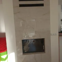 Obudowa kominka wykończona płytami imitującymi marmur z czarnymi wstawkami wentylacyjnymi i czarnym wkładem kominkowym, widoczne przełączniki elektryczne i fragment zabawki dziecięcej.