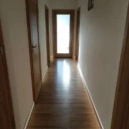 Długi korytarz z drewnianą podłogą i listwami przypodłogowymi z oświetleniem LED, widoczne trzy drewniane drzwi i obrazek na ścianie.