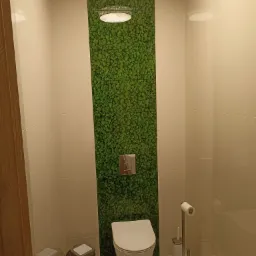 Nowoczesna łazienka z toaletą podwieszaną i pionowym panelem z mchu dekoracyjnego nad nią, oświetlonym punktowo, kremowe płytki na ścianach, widoczny kosz na śmieci i akcesoria toaletowe.