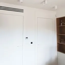 Białe, minimalistyczne skrzydła wbudowane w ścianę, z ukrytymi zawiasami i minimalistycznymi klamkami, z widocznymi włącznikami światła, na podłodze jasny parkiet, oświetlenie punktowe sufitowe...