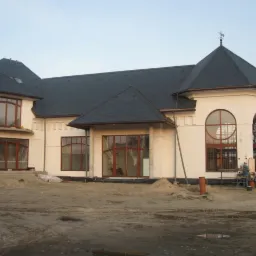 Wznoszony, biały budynek z ciemnym dachem, dużymi oknami w drewnianych ramach i dekoracyjną wieżyczką z wiatrowskazem, na placu budowy z piaskiem i widocznymi pracownikami w oddali.