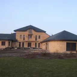 Duży, parterowy dom z dobudówką w kształcie rotundy, wykończony piaskowym kamieniem, kryty ciemną dachówką, z ogrodem w trakcie aranżacji.