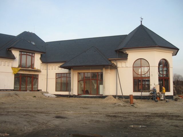 Wznoszony, biały budynek z ciemnym dachem, dużymi oknami w drewnianych ramach i dekoracyjną wieżyczką z wiatrowskazem, na placu budowy z piaskiem i widocznymi pracownikami w oddali.