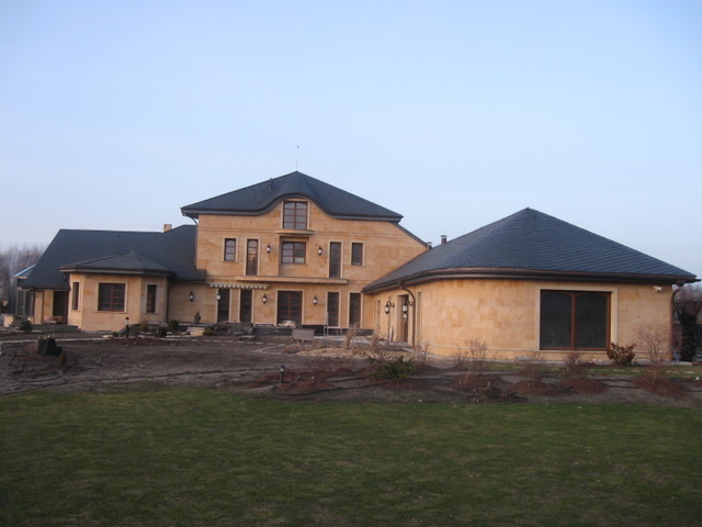 Duży, parterowy dom z dobudówką w kształcie rotundy, wykończony piaskowym kamieniem, kryty ciemną dachówką, z ogrodem w trakcie aranżacji.