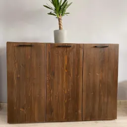 Brązowa szafka z trzema drzwiczkami z widocznym usłojeniem drewna i czarnymi, prostokątnymi uchwytami, na górze doniczka z zieloną rośliną.