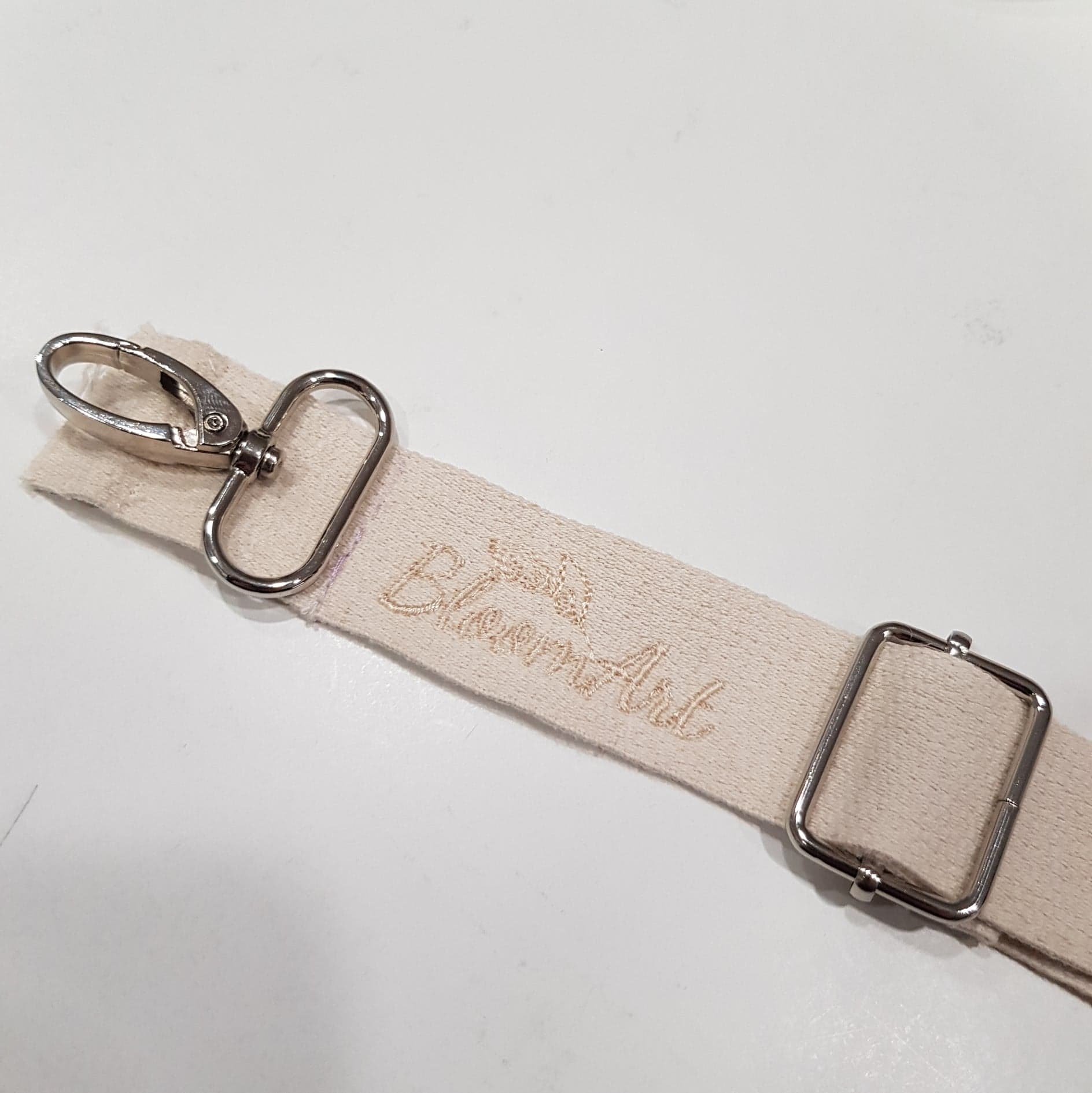 Pasek z beżowej tkaniny z wyhaftowanym napisem 'Bloom Art' i metalowymi elementami mocującymi, leżący na białym tle.