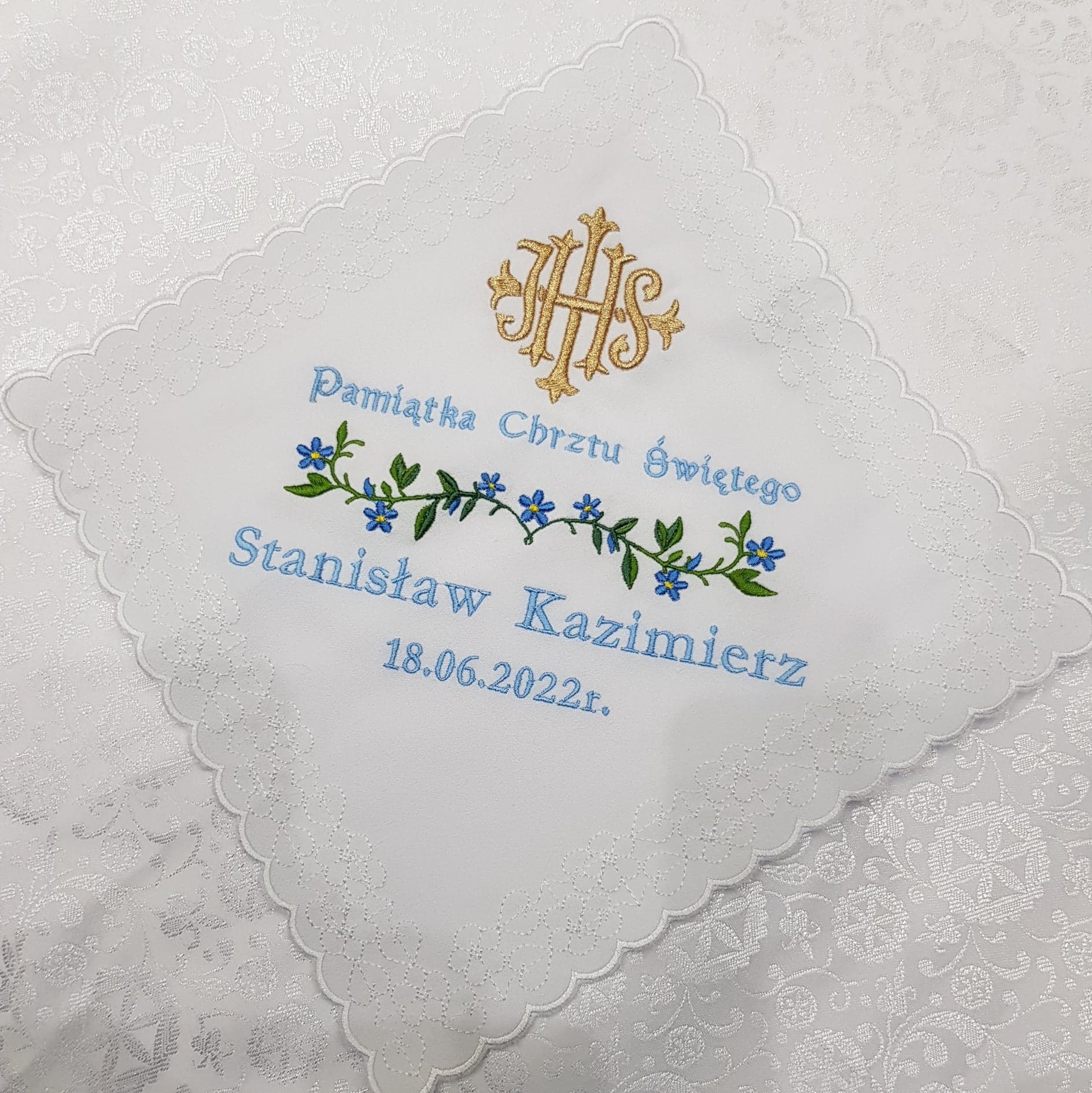 Biała chusteczka z haftowanym złotym monogramem IHS, niebieskim napisem 'Pamiątka Chrztu Świętego', imieniem Stanisław Kazimierz, datą 18.06.2022r. oraz motywem roślinnym z niebieskimi kwiatkami...