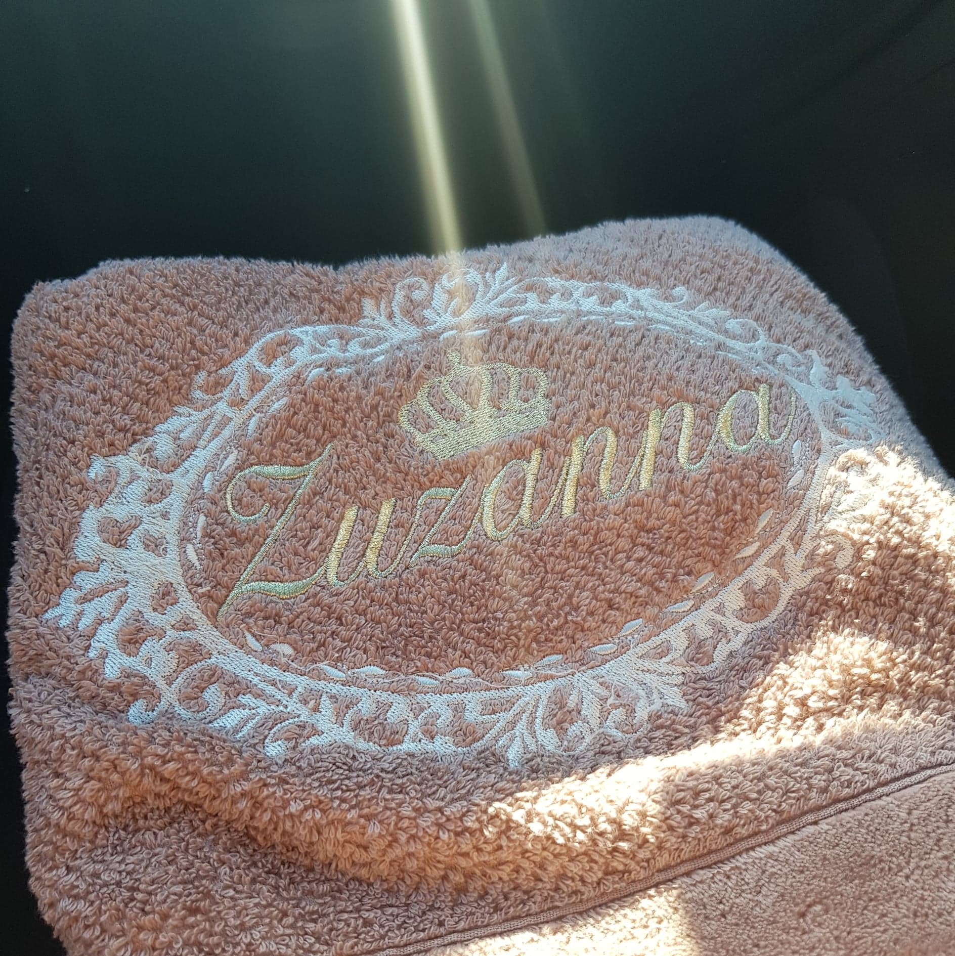 Różowy ręcznik frotte z wyhaftowanym imieniem 'Zuzanna' w ozdobnej ramce i koroną, oświetlony promieniami słońca.