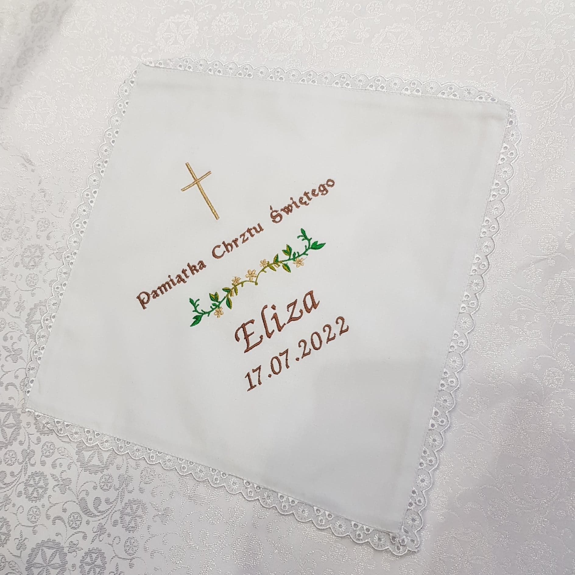 Biała, kwadratowa chusteczka z haftowanym napisem 'Pamiątka Chrztu Świętego', imieniem 'Eliza', datą '17.07.2022' oraz krzyżem i motywem roślinnym, wykończona koronką, leżąca na białym obrusie...