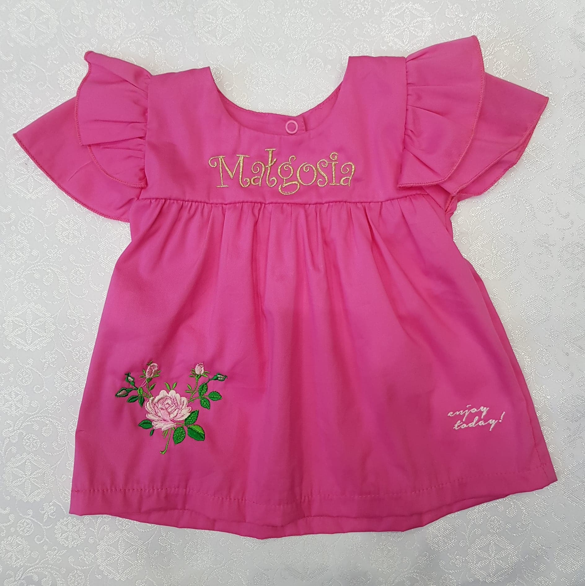 Różowa, bawełniana sukienka dziecięca z haftowanym imieniem 'Małgosia', kwiatowym motywem i napisem 'enjoy today!' na białym, wzorzystym tle.