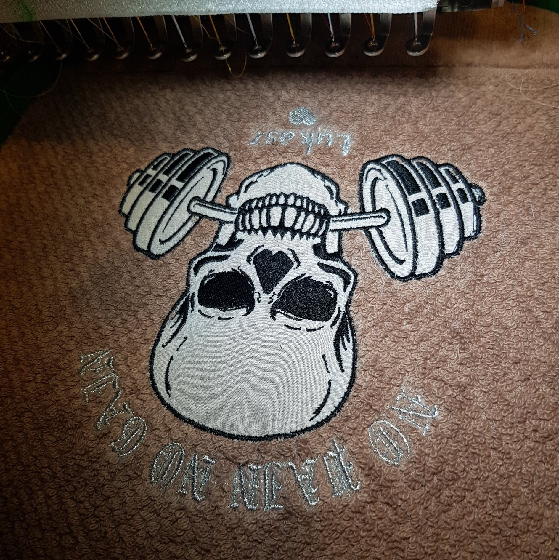 Haftowany na brązowym ręczniku motyw czaszki dźwigającej sztangę, z napisem 'Lukasz' u góry i 'NO PAIN NO GAIN' na dole, wykonany na maszynie do haftowania.