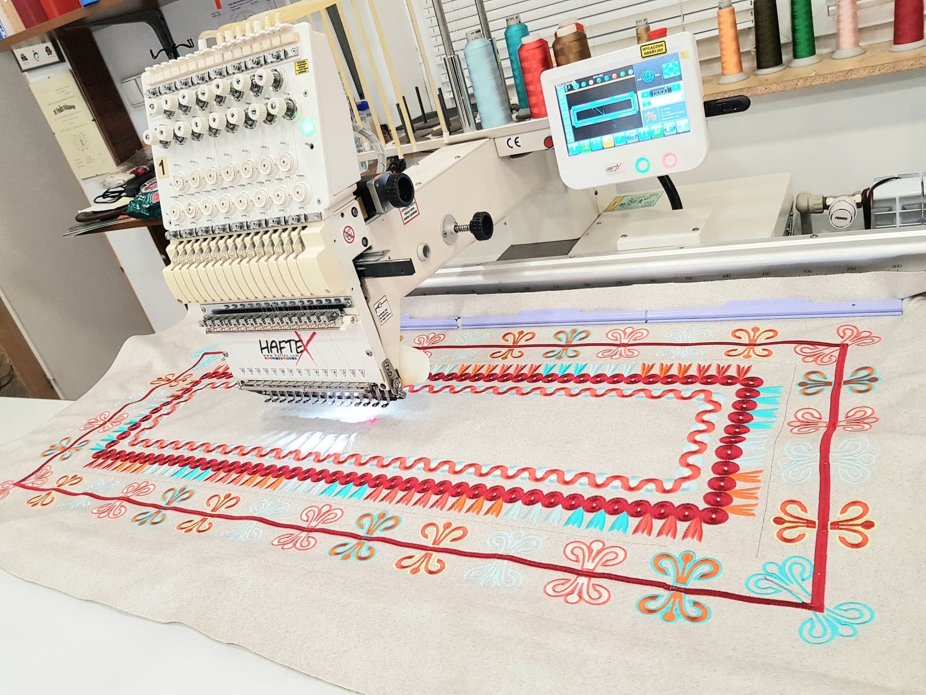 Automatyczna maszyna do szycia haftuje wzór na beżowej tkaninie, widoczne kolorowe nici i ekran sterujący maszyną.