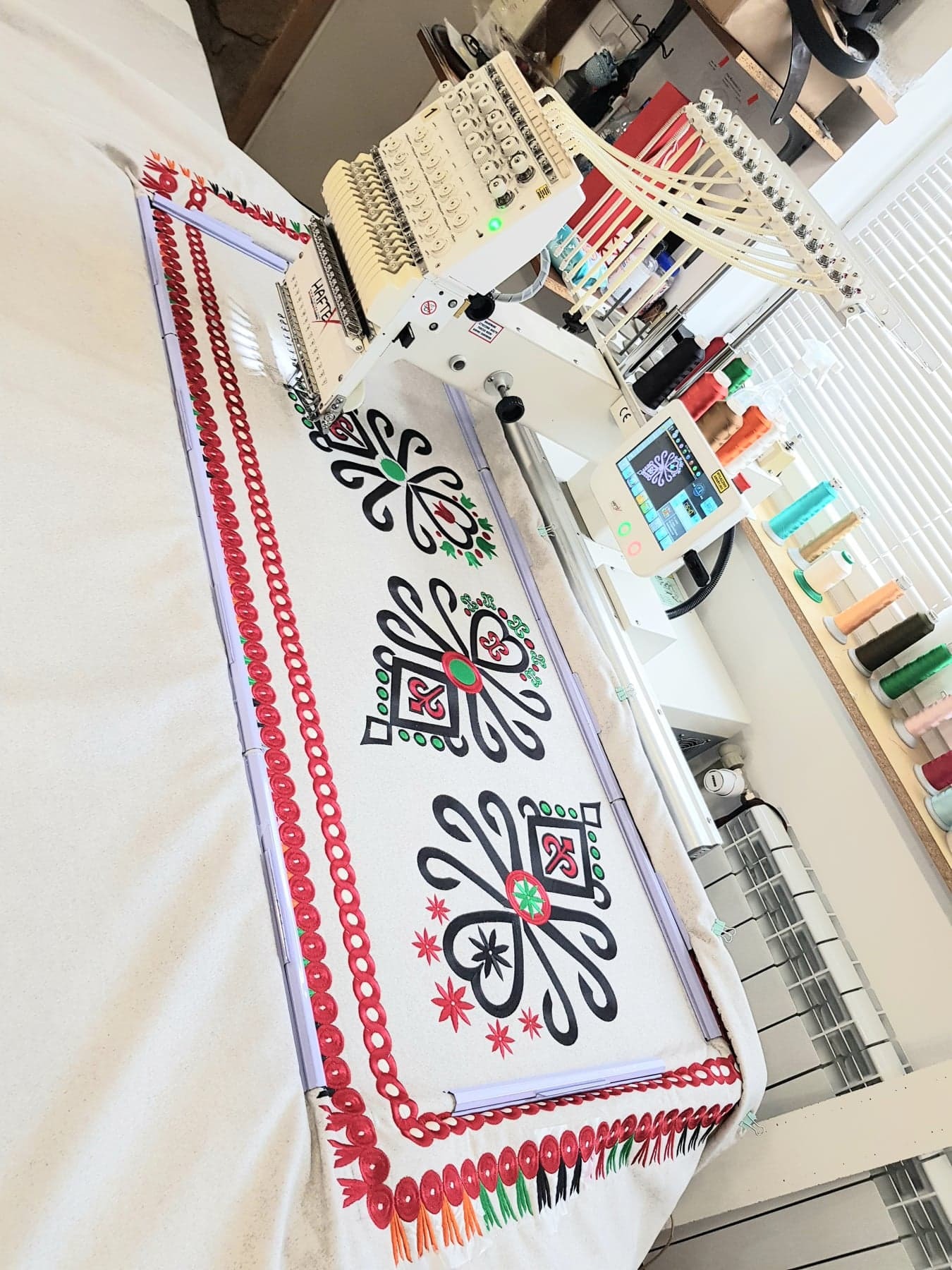 Automatyczna hafciarka w trakcie pracy nad wzorem ludowym na białym materiale, widoczne kolorowe nici i ekran maszyny.