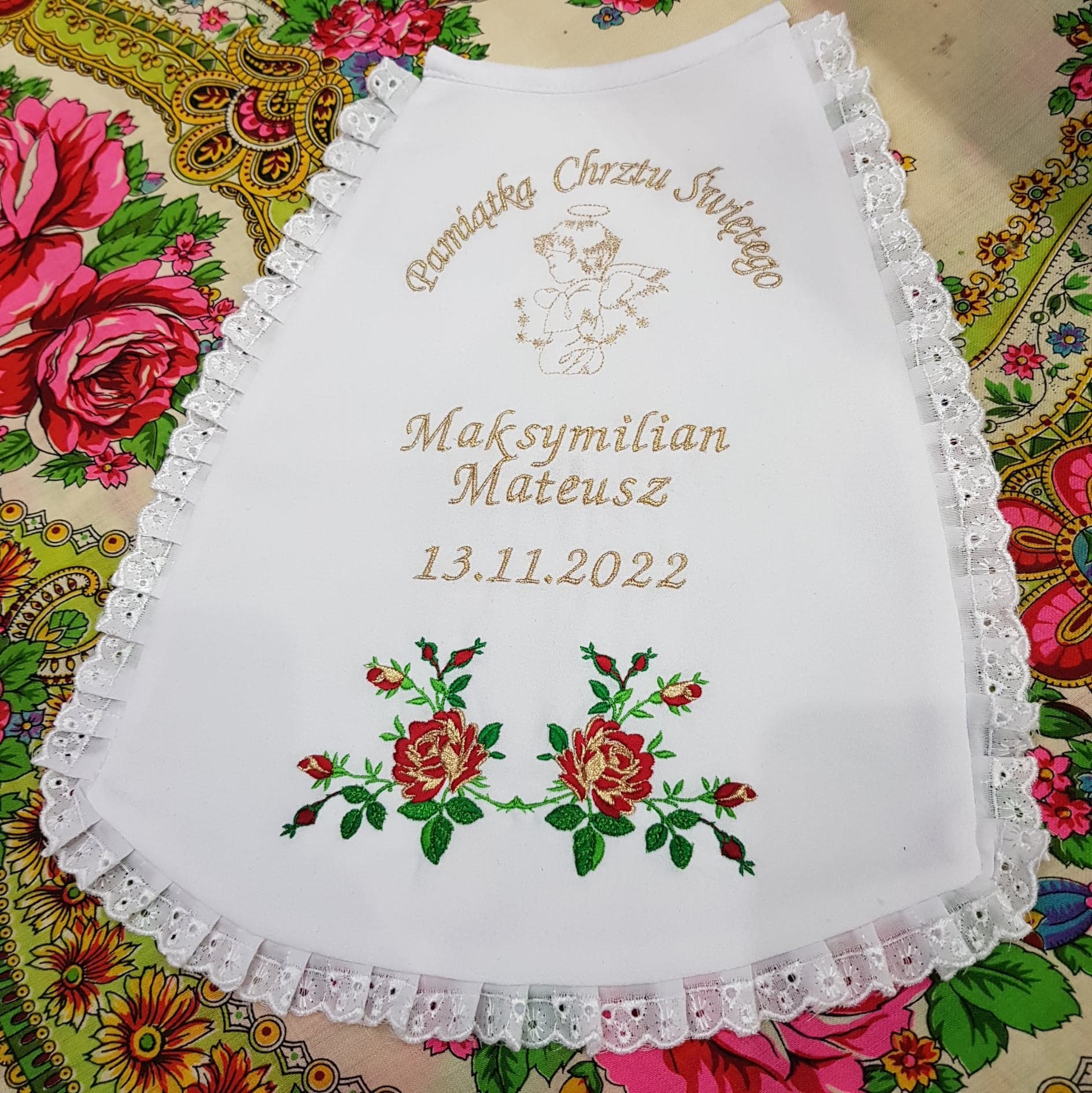 Biała, haftowana pamiątka chrztu z imieniem Maksymilian Mateusz i datą 13.11.2022, ozdobiona koronkową falbanką i motywem haftowanych czerwonych róż, leżąca na kwiecistej tkaninie.