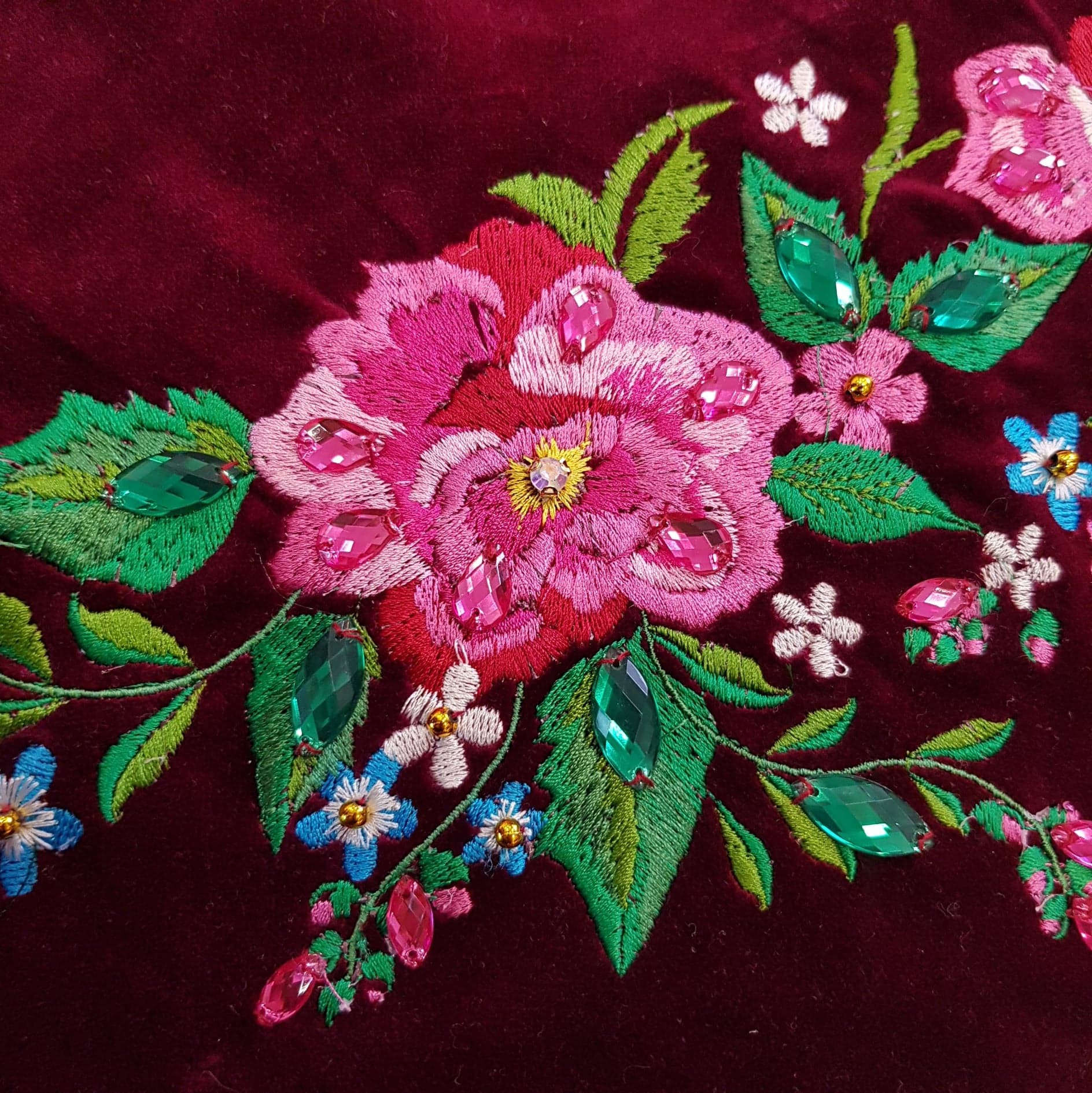 Fragment bordowego weluru z haftowanym, stylizowanym kwiatem w odcieniach różu i zieleni, zdobionym różowymi i zielonymi kryształkami.