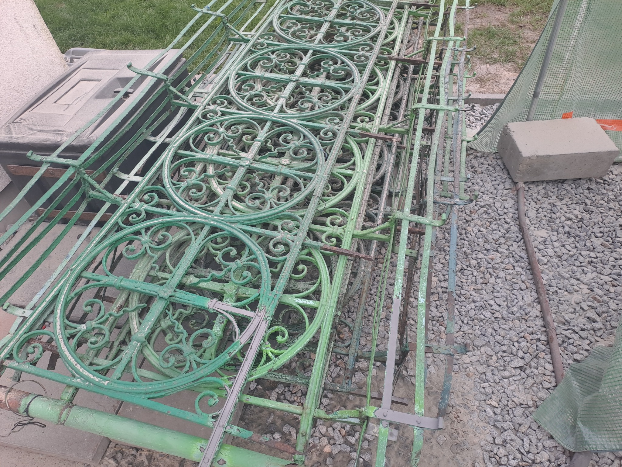 Stos zielonych, metalowych elementów ogrodzenia z dekoracyjnymi okręgami, pokrytych łuszczącą się farbą, składowanych na żwirowym podłożu.