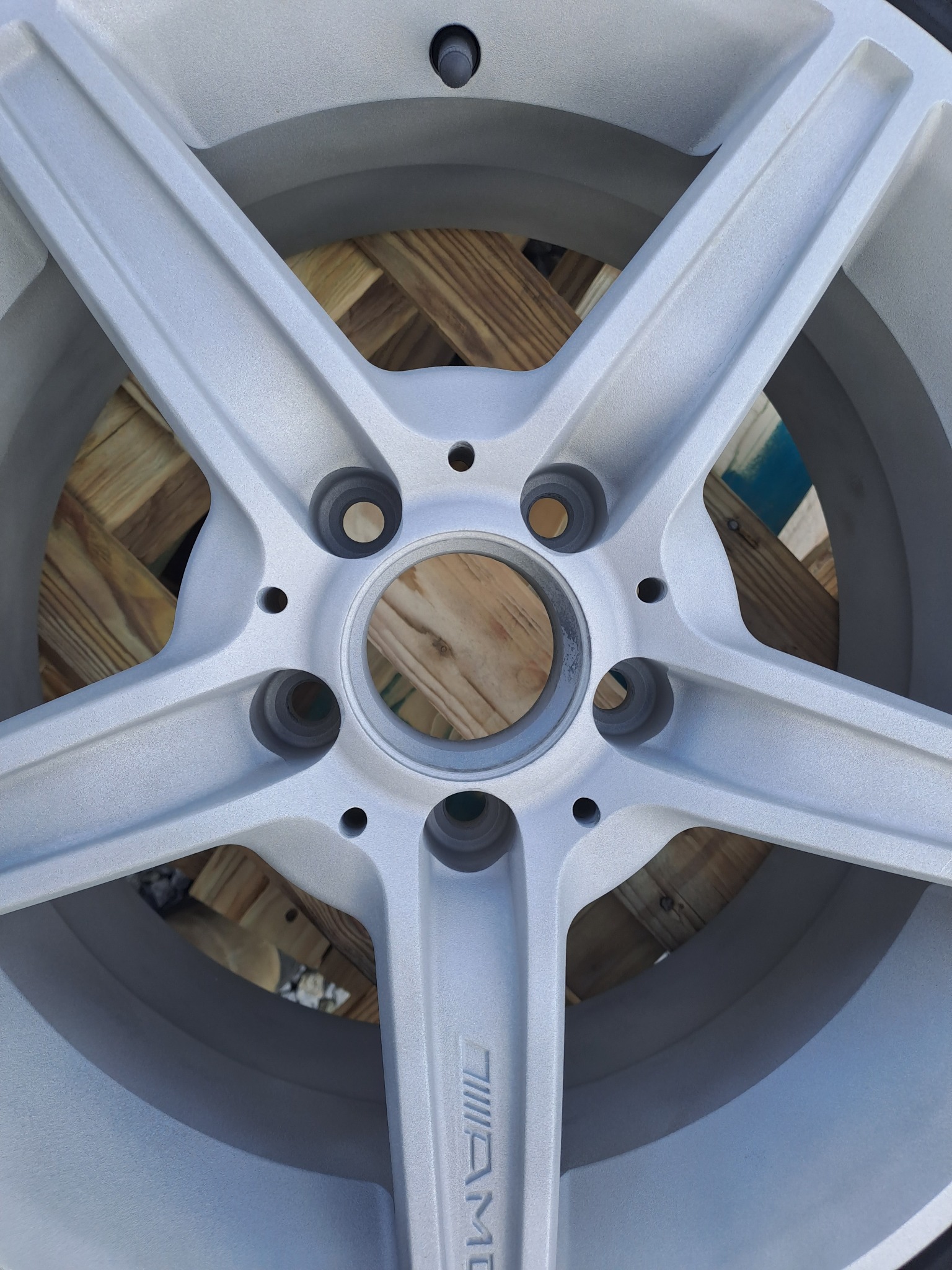 Szara felga aluminiowa AMG po odnowieniu, widoczna faktura powierzchni i detale mocowania na tle drewnianej konstrukcji.