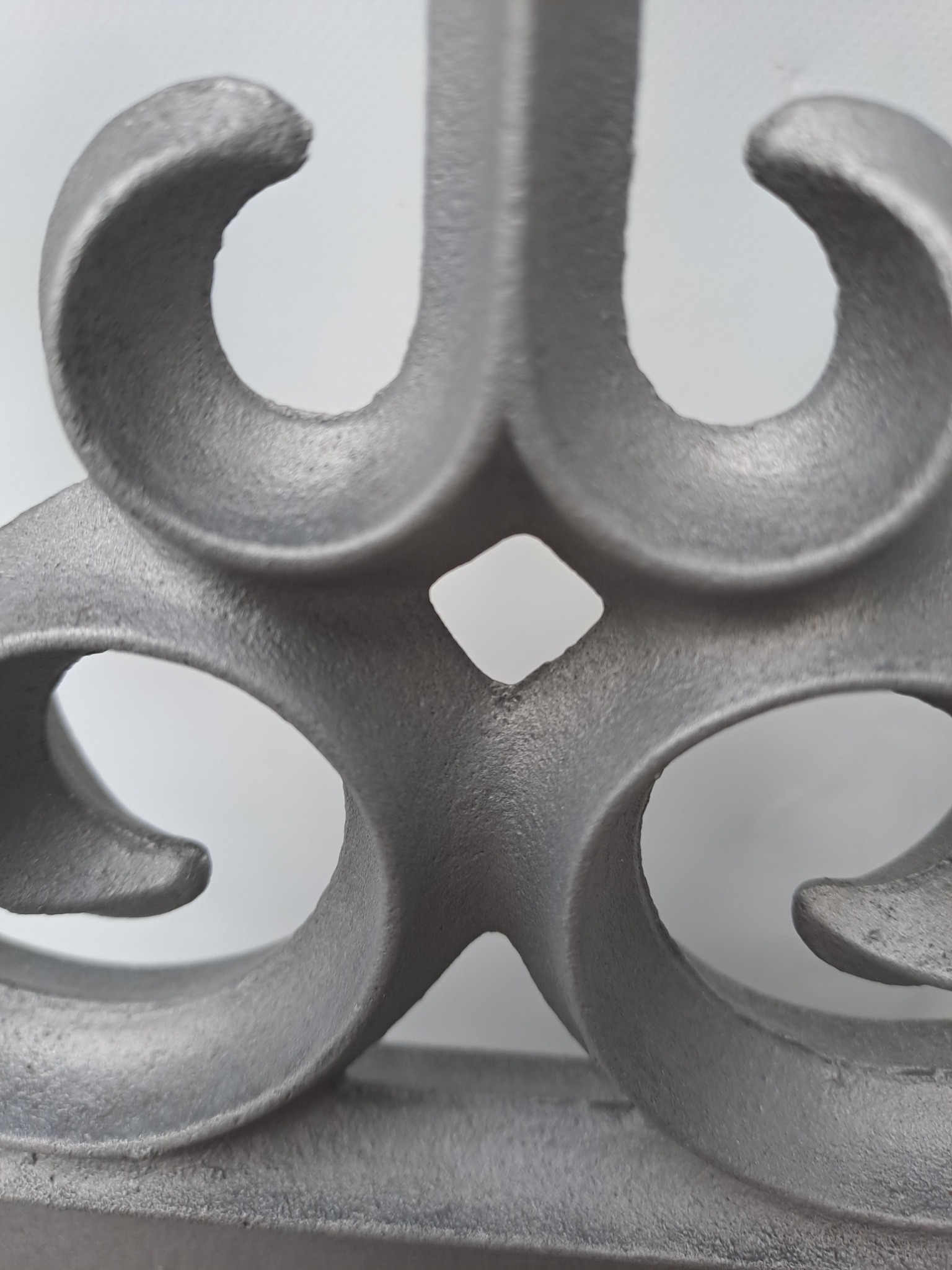 Metalowy, szary ornament z symetrycznymi, zakrzywionymi elementami na jasnym tle, widoczne delikatne nierówności powierzchni.