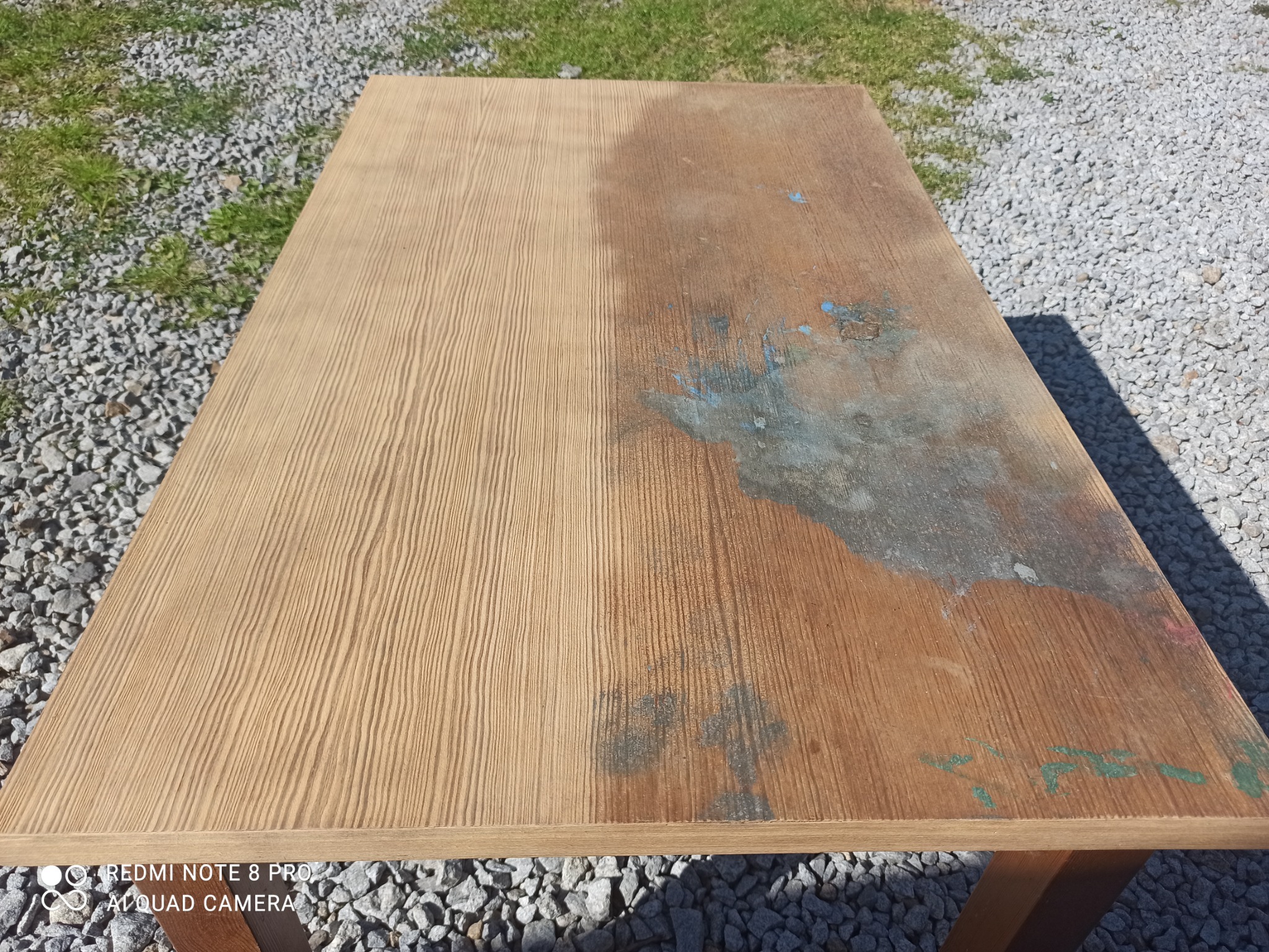Częściowo oczyszczony drewniany blat stołu na zewnątrz, z widocznymi śladami starej farby i zanieczyszczeń.